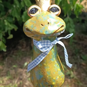Op de afbeelding: Een keramieken kikkerbeeldje met een glanzend geel en blauw gespikkeld glazuur, voorzien van een goudkleurige kroon op het hoofd en een lichtblauw met wit gingham-lint gestrikt in een strik om de nek, met grote, donkere, expressieve ogen.