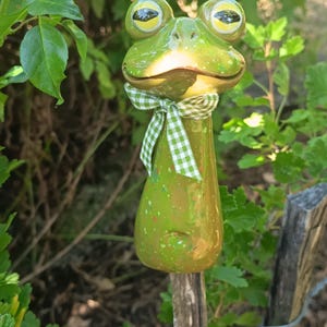 Può includere: Un picchetto da giardino in ceramica verde lucido a forma di rana, con grandi occhi gialli e neri, una bocca larga e un piccolo nastro in tessuto a quadretti verdi e bianchi legato al collo. Il corpo presenta una finitura maculata.