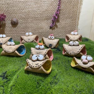 Può includere: Una collezione di fantasiose statuette di cannoli in ceramica fatte a mano con facce dipinte e interni colorati. I cannoli sono disposti su un letto di erba artificiale verde, con uno sfondo neutro di tela di juta.