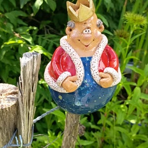 Peut inclure: Une décoration de jardin en céramique en forme de roi rond portant une couronne dorée, une veste rouge à bordure blanche et une base bleue. La figurine a un visage beige clair, de petites oreilles et une expression souriante, fixée sur un poteau.