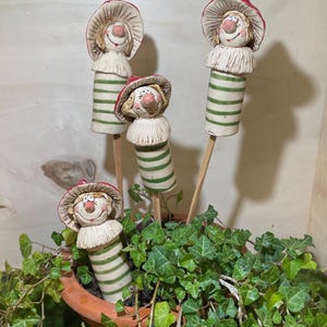 Può includere: Quattro spiritosi picchetti da giardino in ceramica. Ogni picchetto presenta un volto sorridente con un cappello bordato di rosso e un colletto a balze. I corpi cilindrici sono a righe verdi e bianche. I picchetti sono in un vaso di terracotta pieno di edera verde.