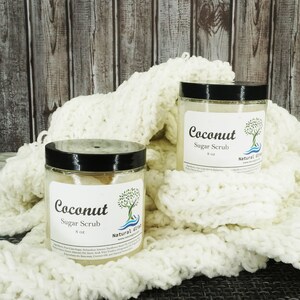 May include: Two glass jars of coconut sugar scrub, each with a black lid and a white label that says "Coconut Sugar Scrub" and "Natural  Slist". The jars are sitting on a white knitted blanket.