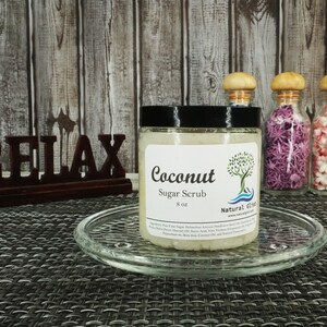 May include: A clear glass jar with a black lid filled with a white sugar scrub. The jar is labeled "Coconut Sugar Scrub 8 oz" and "Natural Gifts". The jar is sitting on a glass plate on a woven placemat. The word "RELAX" is written in brown letters on a wooden sign behind the jar.