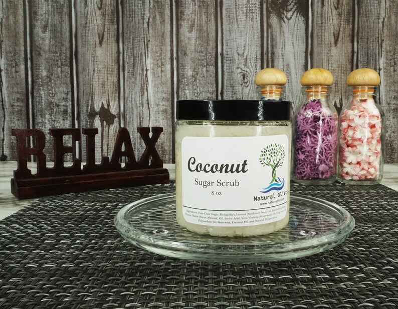 May include: A clear glass jar with a black lid filled with a white sugar scrub. The jar is labeled "Coconut Sugar Scrub 8 oz" and "Natural Gifts". The jar is sitting on a glass plate on a woven placemat. The word "RELAX" is written in brown letters on a wooden sign behind the jar.