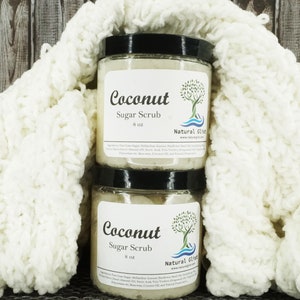 May include: Two glass jars of coconut sugar scrub, each labeled "Coconut Sugar Scrub 8 oz" with a tree and wave design. The jars are sitting on a white knitted blanket.