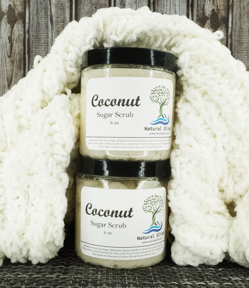 May include: Two glass jars of coconut sugar scrub, each labeled "Coconut Sugar Scrub 8 oz" with a tree and wave design. The jars are sitting on a white knitted blanket.