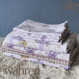 May include: A stack of folded bed linens featuring floral and striped patterns in shades of white, lavender, and purple. The linens are arranged on a white lace surface against a blue textured wall.