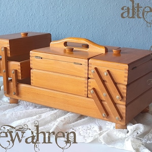 Könnte beinhalten: Ein hellbrauner Nähkasten aus Holz mit ausziehbarem Ziehharmonika-Mechanismus, mehreren Fächern, aufklappbaren Deckeln mit kleinen runden Knöpfen und einem zentralen Tragegriff. Der Kasten ist mit sichtbaren Fingerzinken und Metallscharnieren gefertigt.