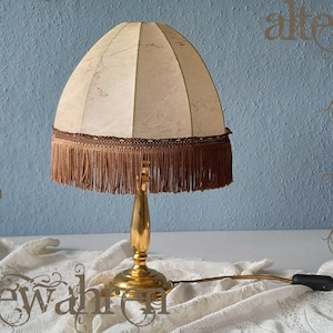 May include: A vintage table lamp with a beige lampshade and brown fringe. The lamp has a gold-coloured base and stem. The cord and plug are visible. The lamp is on a white surface against a blue wall.