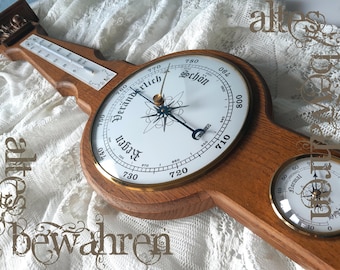 May include: A wooden weather station featuring a central barometer with a white dial, a thermometer at the top, and a hygrometer at the bottom. The barometer dial displays the German text Regen, Veränderlich, and Schön with black hands.