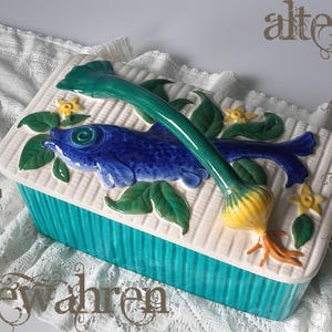 Op de afbeelding: Een rechthoekige keramische doos met deksel, een geribbelde turquoise basis en een crèmekleurig deksel. Het deksel is voorzien van een reliëf van een blauwe vis, groene bladeren, kleine gele bloemen en een handvat in de vorm van een ui.