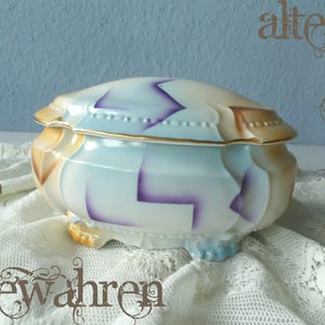 May include: A porcelain lidded box with a glossy finish, featuring a cream base color with light blue and purple geometric accents. The container has a scalloped rim with gold trim and rests on four small, molded feet with a textured surface.