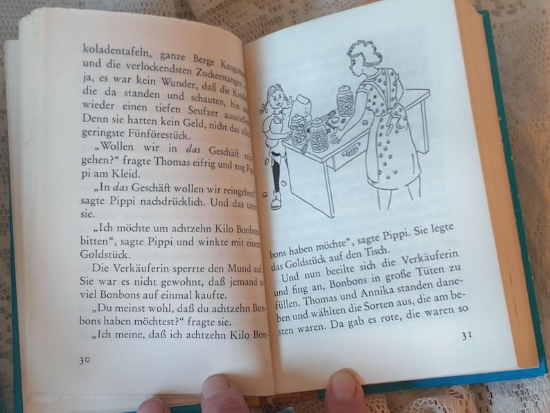 May include: Open book with text and a black and white illustration of a girl and a woman behind a counter. The text is in German. The book has a blue cover.