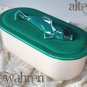 Può includere: Un piatto da portata in ceramica con coperchio verde acqua con una statuina di foca. Il piatto ha una base bianca con scanalature verticali e forma ovale. Il coperchio è verde acqua e la foca è di un colore verde acqua più scuro.