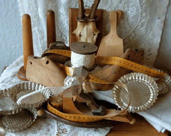 Op de afbeelding: Een verzameling vintage naai- en knutselgereedschap, waaronder een houten spoelhouder, een houten schoenleest, een spoel wit garen, een meetlint en een set glazen taartvormen.