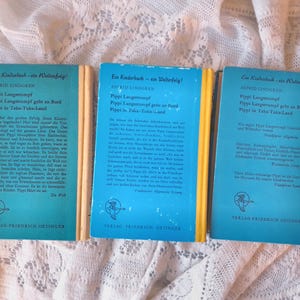 May include: Three vintage children's books with blue covers and yellow spines are displayed. The titles include "Pippi Longstocking" in German. The publisher's logo is a bird. The books are on a white lace surface.