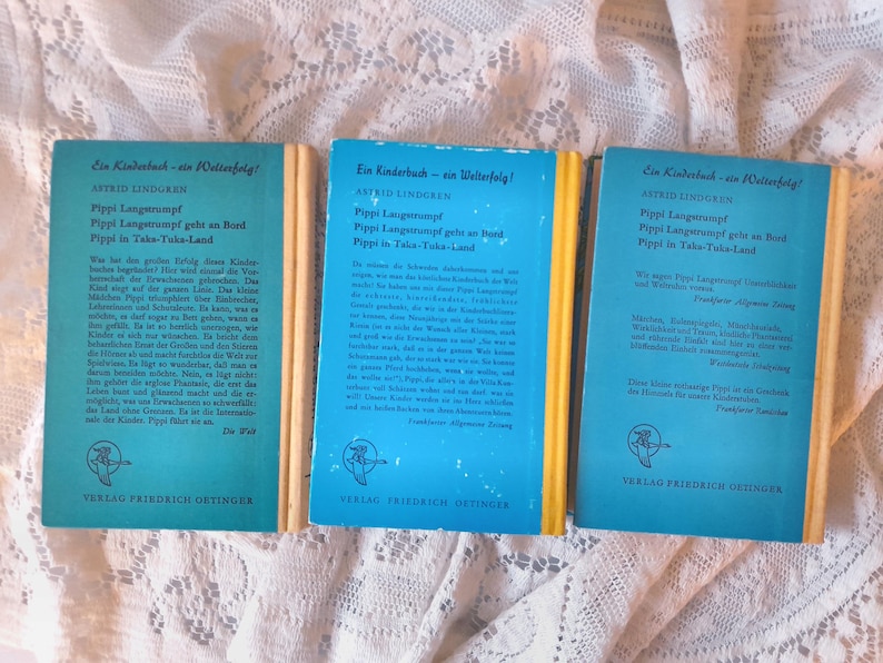 May include: Three vintage children's books with blue covers and yellow spines are displayed. The titles include "Pippi Longstocking" in German. The publisher's logo is a bird. The books are on a white lace surface.
