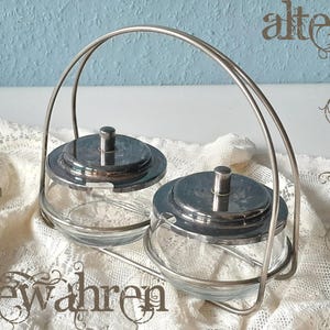 Könnte beinhalten: Ein Vintage-Zuckerstreuer-Set aus Glas und Metall. Das Set besteht aus zwei durchsichtigen Glasschalen mit silberfarbenen Deckeln und einem passenden Griff. Die Schalen stehen auf einer weißen Spitzentischdecke vor einem hellblauen Hintergrund.
