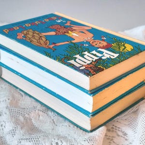 May include: Three vintage hardcover books stacked on top of each other. The top book has a blue cover with an illustration of a person and the title "Pippi in a-a-u-ka-land". The book edges are yellow and the pages are white.