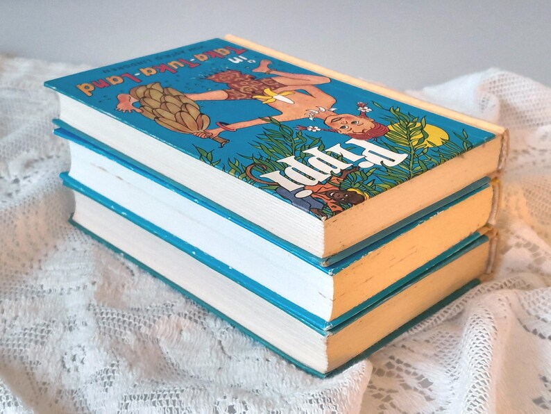 May include: Three vintage hardcover books stacked on top of each other. The top book has a blue cover with an illustration of a person and the title "Pippi in a-a-u-ka-land". The book edges are yellow and the pages are white.