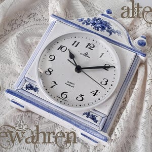 Op de afbeelding: Een witte en blauwe keramische klok met een quartz uurwerk. De wijzerplaat heeft zwarte cijfers en wijzers, met de merknaam "DUGENA" zichtbaar. Bloemenontwerpen en blauwe accenten versieren de lijst van de klok. De klok staat op een wit kanten oppervlak.