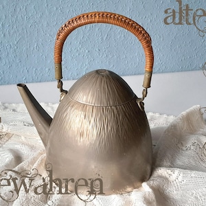 May include: A vintage silver-toned metal teapot with a woven handle. The teapot has a rounded body and a long spout. The handle is made of woven brown material. The image also contains the text "altes Bewahren".