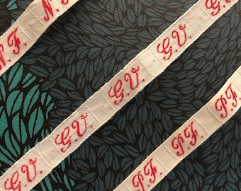 May include: Three white fabric ribbons with red embroidered text. The text on the ribbons reads "N.F.T." and "G.V."