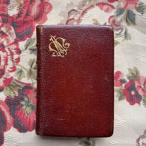 May include: A small, rectangular, leather-bound book in a deep reddish-brown colour. The cover has a gold monogram in an ornate script. The book rests on floral patterned fabric.