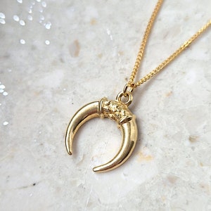 May include: A gold-colored necklace featuring a crescent moon pendant. The pendant has a textured surface and is suspended from a delicate chain. The necklace is displayed on a white and gray marble surface.
