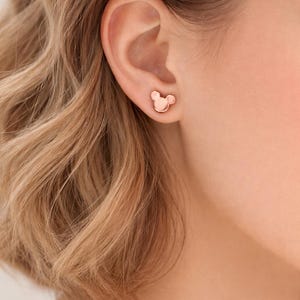 Pode incluir: Brincos de bot&atilde;o em ouro rosa em forma de cabe&ccedil;a de rato. Os brincos s&atilde;o pequenos e simples, com um acabamento liso e polido. S&atilde;o mostrados numa orelha, com o cabelo da pessoa ao fundo.