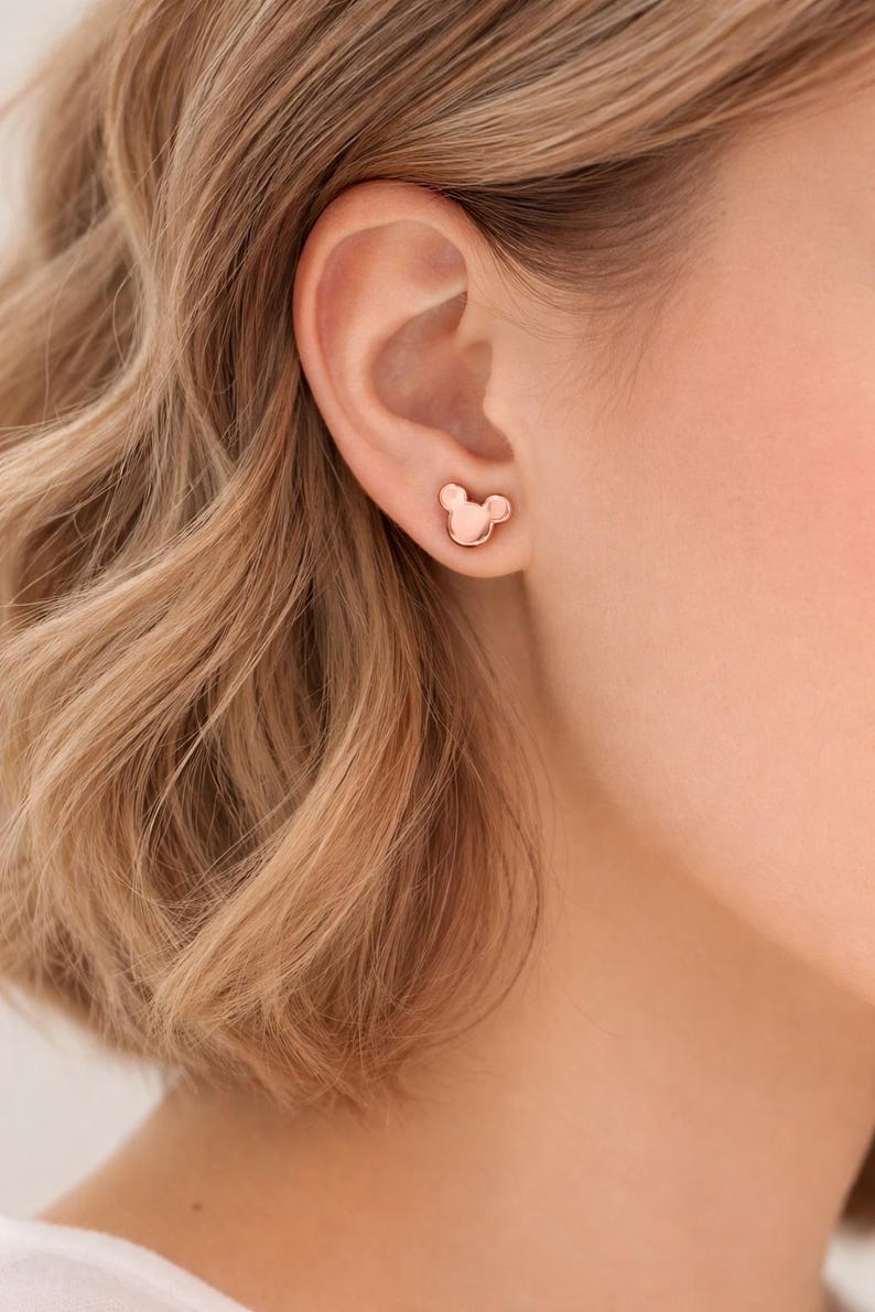 Pode incluir: Brincos de bot&atilde;o em ouro rosa em forma de cabe&ccedil;a de rato. Os brincos s&atilde;o pequenos e simples, com um acabamento liso e polido. S&atilde;o mostrados numa orelha, com o cabelo da pessoa ao fundo.