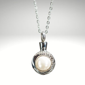 May include: A silver-toned necklace with a round pendant. The pendant features a large, white pearl surrounded by a ring of small, clear crystals. The necklace chain is delicate and also silver-toned.
