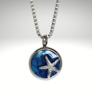 Pode incluir: Um colar de corrente de prata com um pingente redondo que apresenta um design de concha azul e branca com uma estrela do mar prateada no topo.