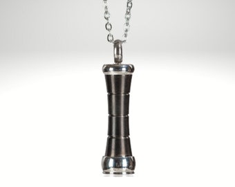 May include: A silver chain necklace with a black and silver cylindrical pendant. The pendant has a textured, layered design.