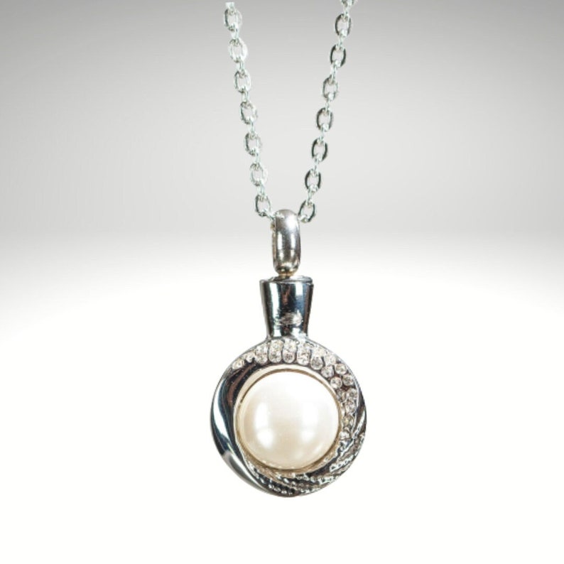 May include: A silver-toned necklace with a round pendant. The pendant features a large, white pearl surrounded by a ring of small, clear crystals. The necklace chain is delicate and also silver-toned.