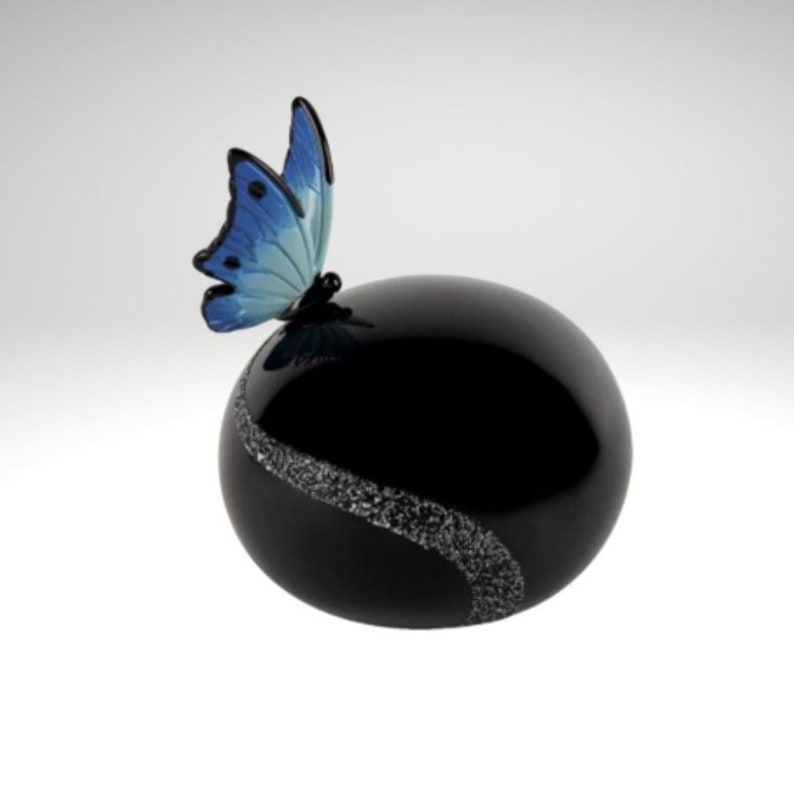 May include: A black glass urn with a silver swirl design and a blue butterfly on top. The urn is a decorative container for ashes.