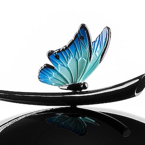 Puede incluir: Una tapa de cer&aacute;mica negra con un dise&ntilde;o de mariposa azul y verde. La mariposa est&aacute; posada sobre una manija curva negra.