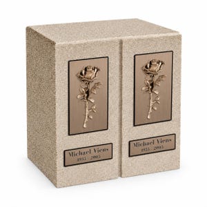 May include: A rectangular beige stone companion urn featuring two bronze-toned plaques, each displaying a raised rose relief. Below each rose, a rectangular bronze nameplate is engraved with the text Michael Viens 1935 - 2005 in black lettering.