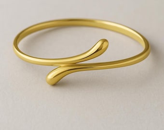 May include: A gold-colored, open cuff bracelet with a minimalist design. The bracelet features a slender band that curves and overlaps, with rounded, teardrop-shaped ends. The jewelry is set against a neutral background.