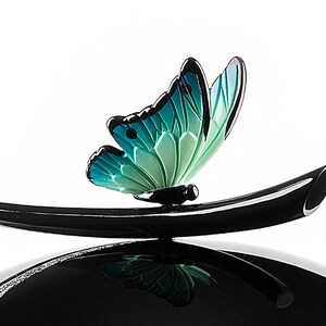 Puede incluir: Una figurita de mariposa negra y verde posada sobre una base negra curva. La mariposa tiene un degradado de color turquesa y verde.
