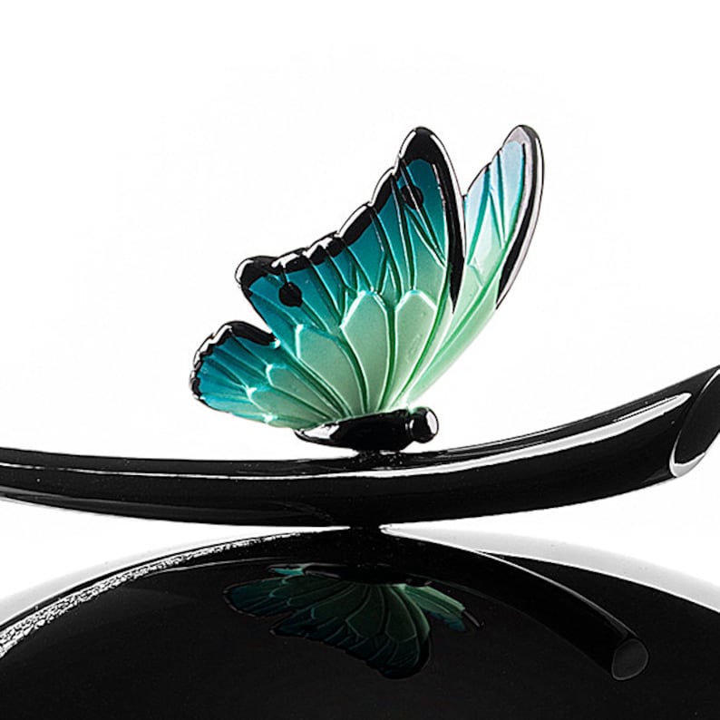 Puede incluir: Una figurita de mariposa negra y verde posada sobre una base negra curva. La mariposa tiene un degradado de color turquesa y verde.