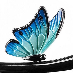 May include: A blue and green butterfly with black accents is perched on a black metal branch. The butterfly has a gradient of color from dark blue to light green.