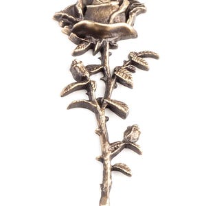 May include: A metallic bronze-toned rose brooch featuring a single blooming flower at the top, a thorny stem, and two smaller rosebuds extending from the sides. The surface has a textured, antique finish with dark highlights in the crevices.