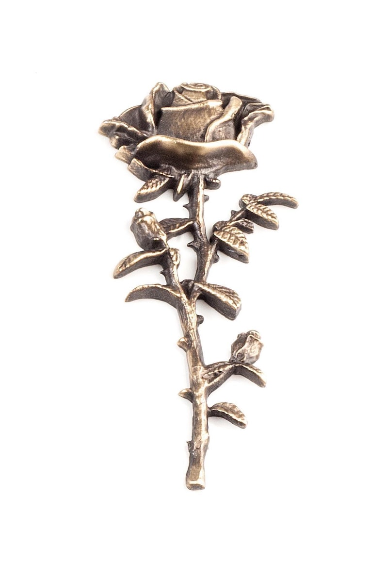 May include: A metallic bronze-toned rose brooch featuring a single blooming flower at the top, a thorny stem, and two smaller rosebuds extending from the sides. The surface has a textured, antique finish with dark highlights in the crevices.