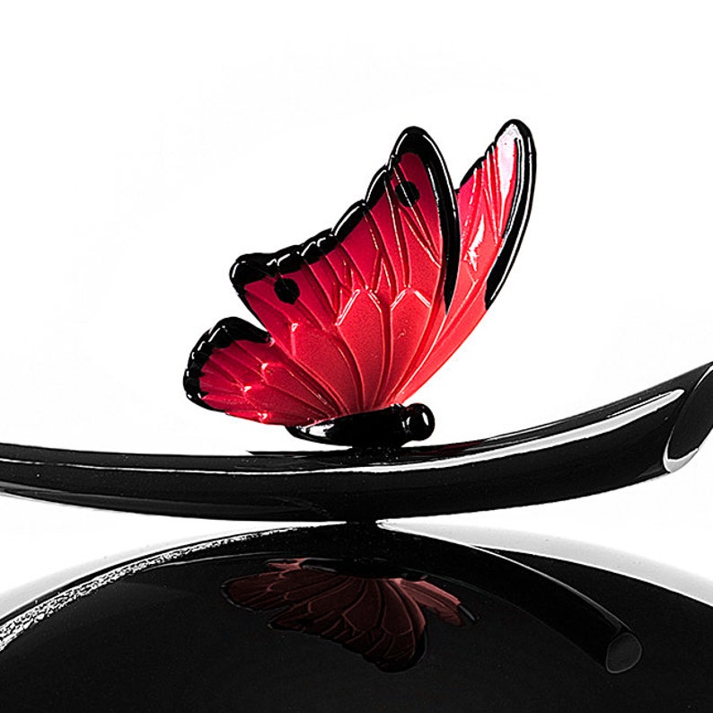 Puede incluir: Un adorno de mariposa de vidrio negro y rojo con un tallo negro. La mariposa tiene un cuerpo rojo y alas negras.