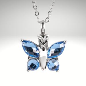 May include: A silver butterfly pendant necklace with blue crystal accents. The pendant is a cremation urn for holding ashes.