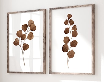 May include: Two framed prints of brown pressed leaves on a white background, hanging on a white wall above a wooden bench with a white throw blanket and a book on top.