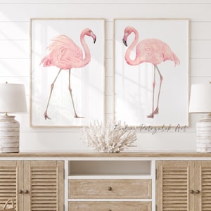 May include: Two framed vertical wall art prints featuring watercolor illustrations of pink flamingos on a white background. The prints are displayed above a wooden media console with louvered cabinet doors and two central drawers.