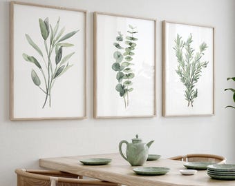 May include: Three framed watercolour prints of different types of greenery, hanging on a white wall above a wooden dining table with four chairs. The table is set with plates and a teapot.