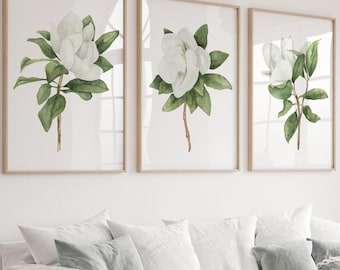 Könnte beinhalten: Ein Set aus drei gerahmten Wandbildern mit Aquarellillustrationen von weißen Magnolienblüten mit grünen Blättern auf weißem Hintergrund, präsentiert in hellen Holzrahmen über einem weißen Sofa in einem Wohnzimmer.